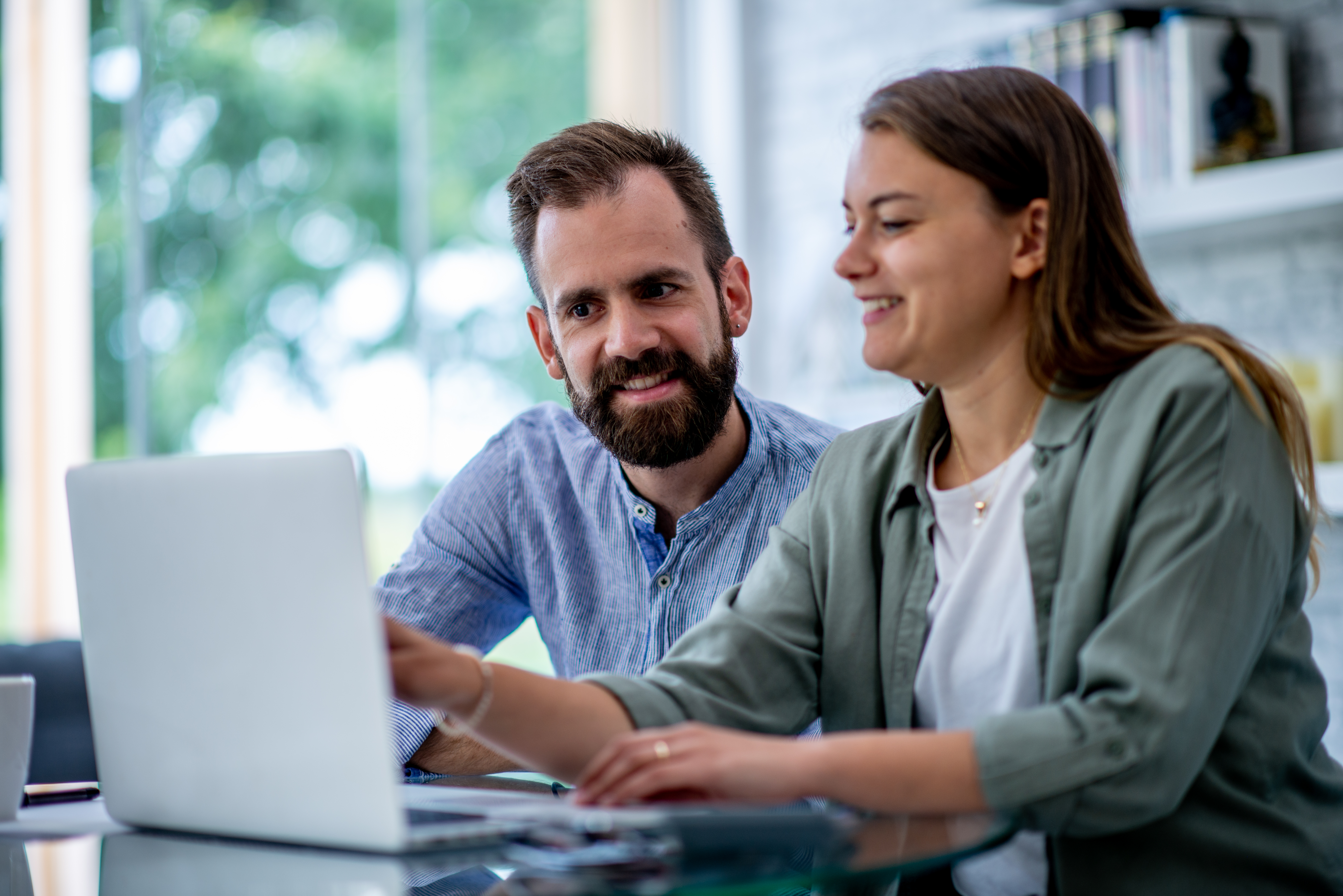 A young couple sit together in their home office as they work through paying bills together. They are both dressed casually, have a laptop open in front of them and appear happy.